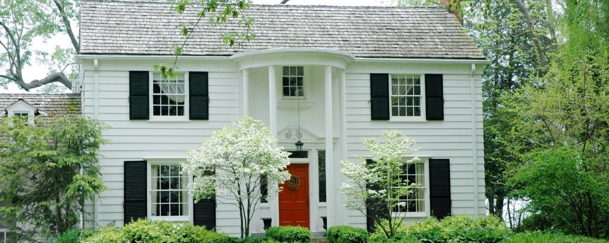 White formal house with siding, black shutters and bright green, manicured lawn and garden