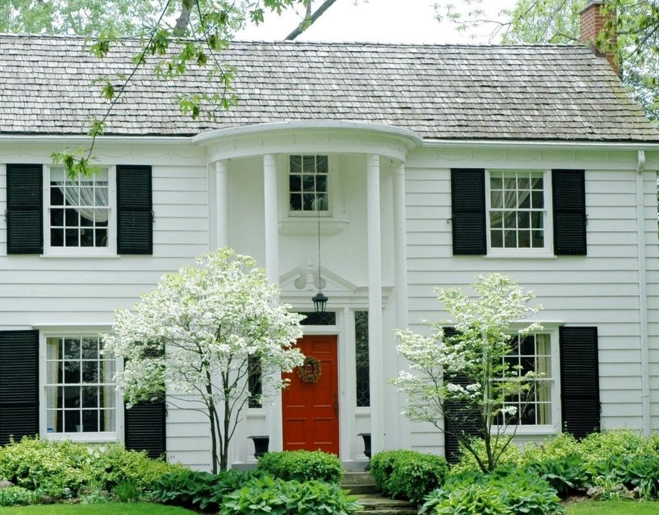 White formal house with siding, black shutters and bright green, manicured lawn and garden