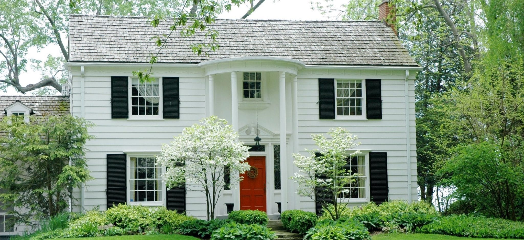 White formal house with siding, black shutters and bright green, manicured lawn and garden