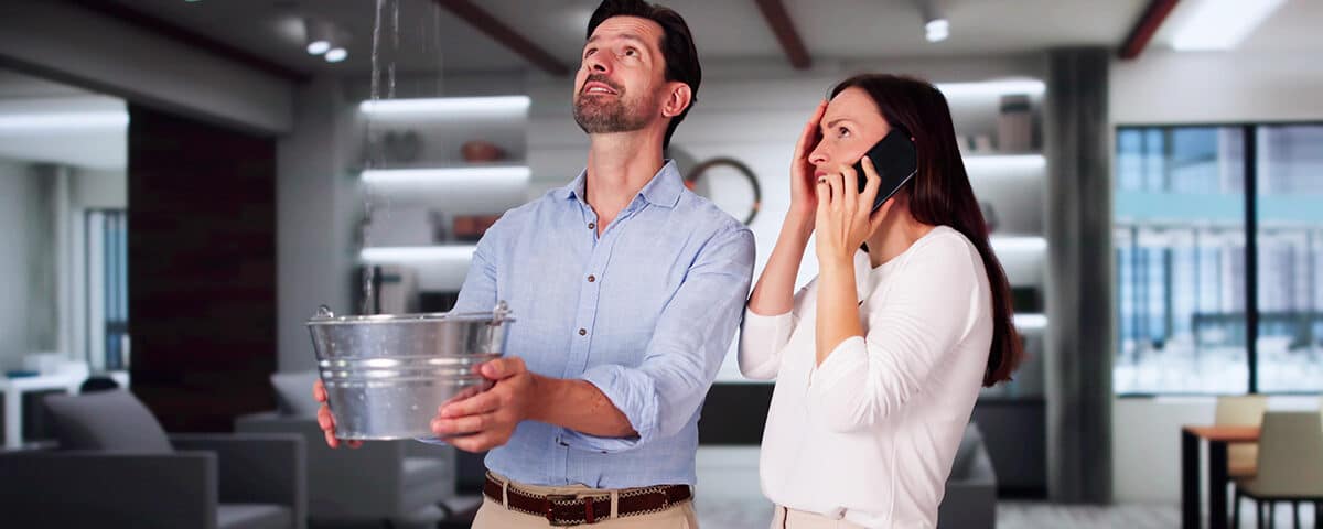 Man catching water leaking from a ceiling in a bucket while a woman makes a call on a mobile phone