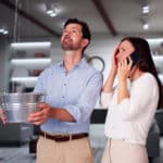 Man catching water leaking from a ceiling in a bucket while a woman makes a call on a mobile phone