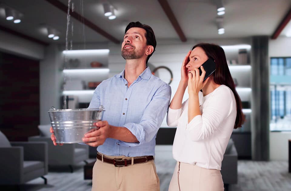 Man catching water leaking from a ceiling in a bucket while a woman makes a call on a mobile phone